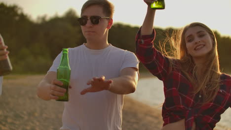 Loving-couple-celebrate-the-end-of-the-semester-with-beer-and-pop-music-on-the-beach-with-their-friends.-They-are-dancing-on-the-open-air-party-at-sunset-in-summer-evening.